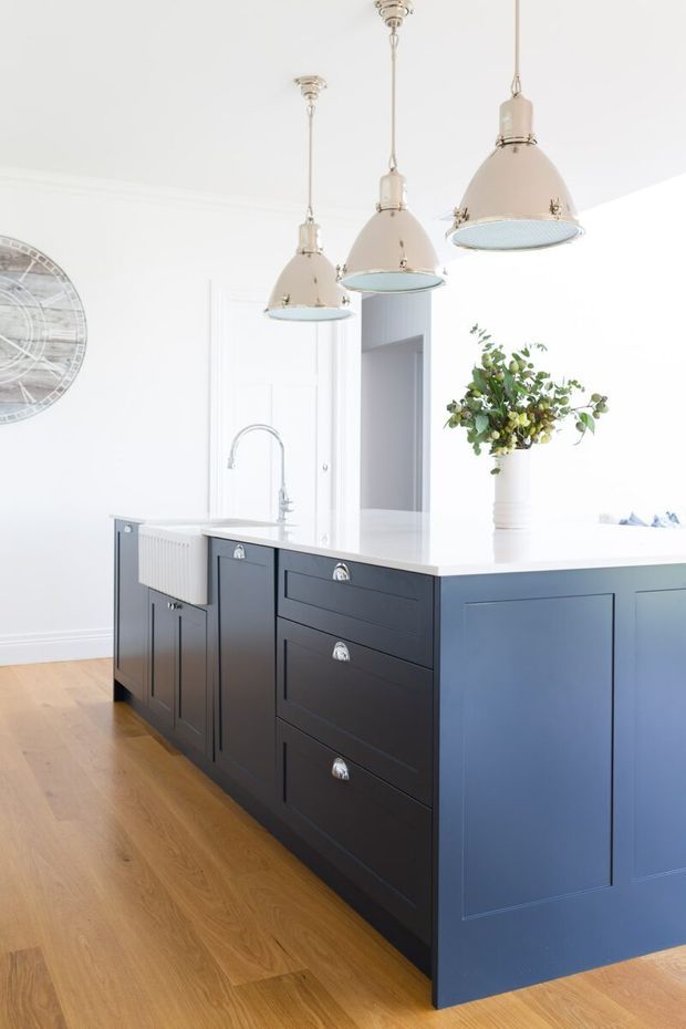 Bright, refined kitchen interior centred on a large dark navy-blue island with shaker-style drawer and door cabinetry, a white benchtop, and an integrated sink with chrome gooseneck tap. Three oversized cream pendant lights hang above the island. Pale timber flooring, white walls and panelling, a floral arrangement, and a large wall clock complete the space.