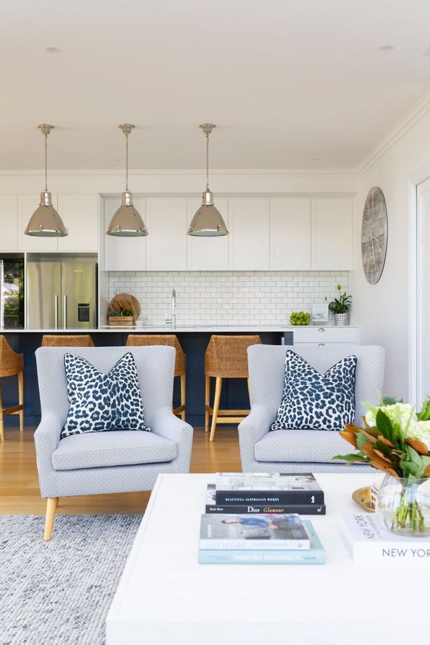 Bright, refined open-plan living and kitchen interior. Two light upholstered armchairs with patterned cushions face a white coffee table, while a compact kitchen sits behind them with white paneled cabinetry, white subway-tile splashback, stainless-steel appliances, a dark island, woven bar stools, and three pendant lights. Warm timber flooring, pale walls, classic ceiling cornicing, and curated accessories create an airy contemporary-classic atmosphere.