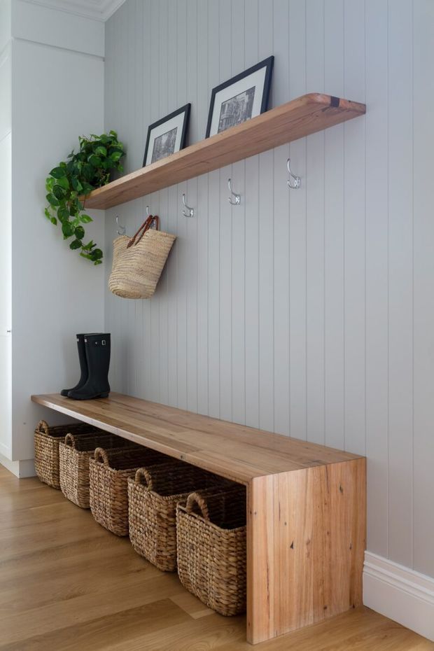 A bright, refined entryway with a built-in timber bench and open wicker basket storage beneath. The wall is clad in vertical pale grey panelling, with a matching timber display shelf, metal hooks, framed artwork, a trailing plant and a pair of boots. Warm timber flooring and white architectural trim complete the welcoming, organised space.