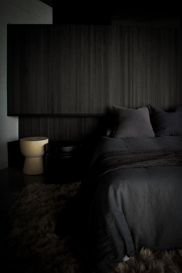 A dark, moody bedroom featuring a low bed dressed in charcoal-grey bedding, a deep vertical timber-panelled feature wall, dark bedside furniture, and a warm cream sculptural bedside lamp. Textural shaggy rugs and layered bedding add softness and contrast to the predominantly black and charcoal palette.