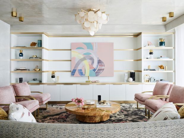 Bright, refined living room with a full-width built-in shelving and cabinetry wall, displaying decorative objects around a large abstract artwork. The room includes a curved textured sofa in the foreground, paired blush-pink armchairs, a sculptural round timber coffee table, patterned area rug, and a statement clustered pendant light. White and pale neutral finishes are accented with brass detailing and soft pink furnishings.