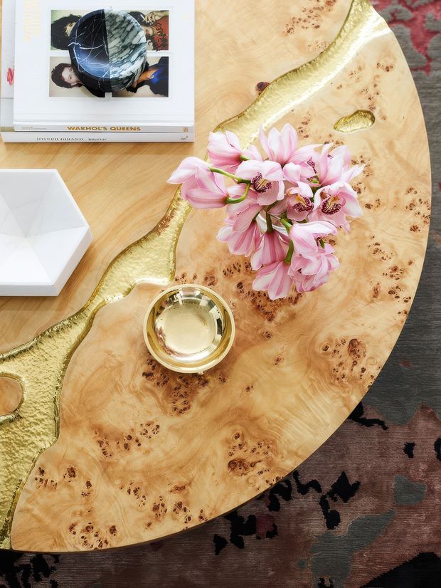 Top-down view of a round live-edge timber coffee table with highly figured burl grain and metallic gold resin-filled natural edges. The table is styled with a gold-toned bowl, pink flowers, a white geometric object, and art books, over a dark patterned rug.