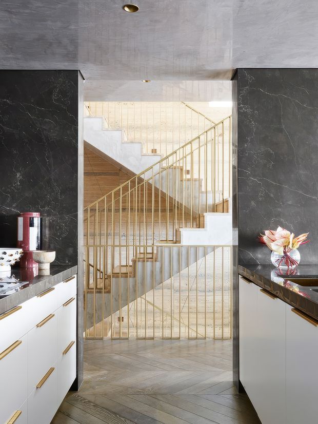 A refined kitchen interior looking toward a sculptural multi-flight staircase. The kitchen has white flat-panel cabinetry with brass pulls, dark veined stone splashbacks and benchtops, and a pale timber floor laid in a herringbone pattern. The staircase features light stone or concrete treads and slender brass-toned vertical balusters, with a bright double-height stair hall beyond. The palette is high-contrast and luxurious, combining black stone, white cabinetry, pale timber, and metallic detailing.