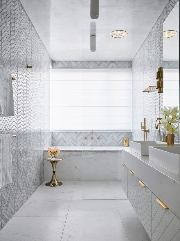 A bright, narrow contemporary bathroom clad in pale grey-white chevron-patterned wall tiles and large marble-look floor tiles. The room features a freestanding bathtub beneath a wide window with a white blind, a floating double vanity with geometric drawer fronts and brass hardware, wall-mounted brass fixtures, a rainfall shower, and a mirrored wall. Gold accents and a small floral arrangement add a refined decorative touch.