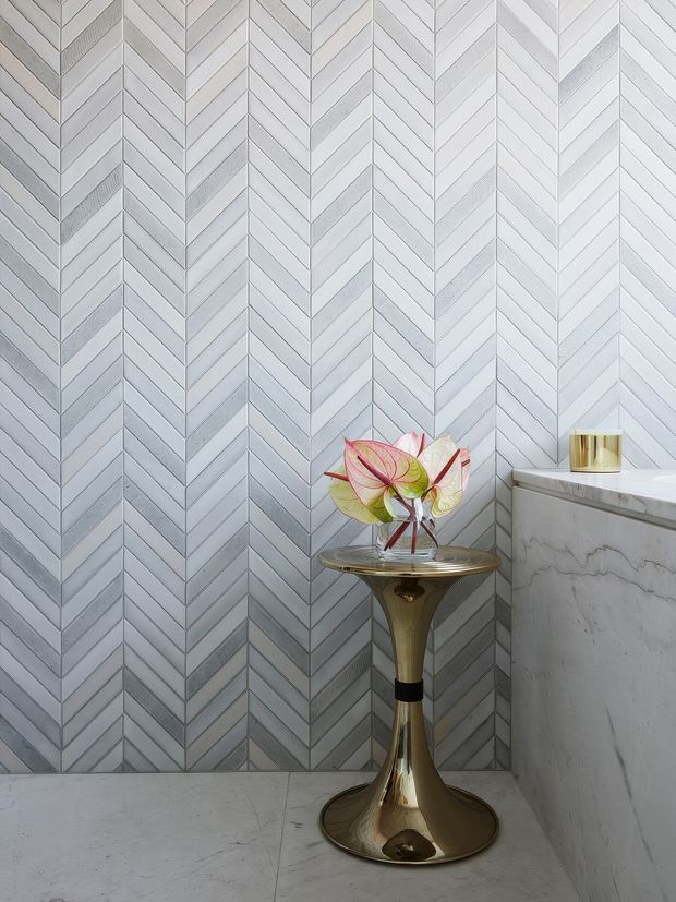 Close-up interior detail of a refined bathroom corner featuring a pale grey and white chevron-patterned wall tile, a matching light stone or marble countertop and side panel, and a sculptural metallic gold pedestal table holding a glass vase of flowers. A small gold-toned candle or vessel sits on the countertop. The composition is bright, polished, and decorative.