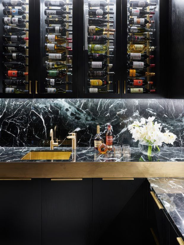 A luxurious dark home bar or kitchen beverage station featuring black cabinetry with brass handles, glass-front wine storage filled with bottles, a gold-toned sink and tap, a dramatic black marble backsplash and countertop, and a timber-toned drawer front. Decorative spirits, glasses, and white flowers sit on the counter.