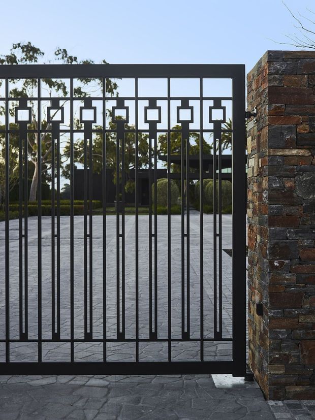 Exterior view of a decorative black metal pedestrian gate set within a dark multicoloured brick masonry wall. The gate features geometric square motifs and slender vertical bars, opening onto a paved driveway or courtyard with landscaped hedges and trees beyond.