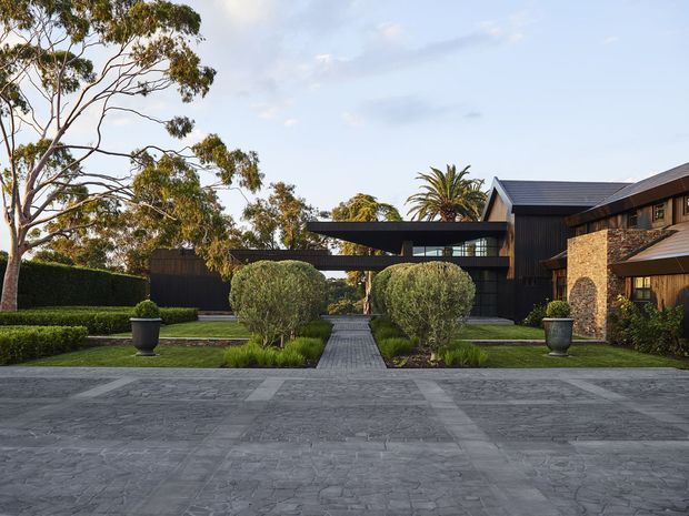 A landscaped approach to a large contemporary residence, with a broad grey paved forecourt leading along a central path through manicured hedges, clipped topiary, lawn and large planters. The house combines dark vertical cladding, extensive glazing, projecting flat-roofed forms and a pitched roof, with a prominent warm stone wall on the right. Mature eucalyptus and palm trees frame the property.