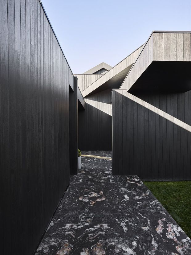 A dramatic exterior architectural walkway between angular, dark vertically clad building volumes. The path is finished in a richly patterned dark stone or concrete surface, with sharp geometric rooflines, projecting forms, and strong light-and-shadow contrasts. A small strip of lawn is visible at the edge beneath a clear sky.