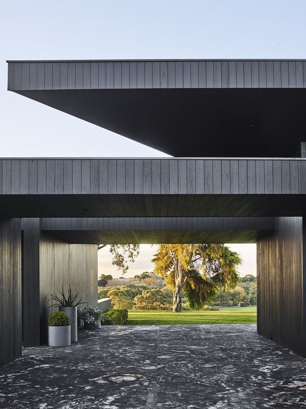 A dramatic contemporary exterior featuring stacked, cantilevered dark volumes clad in vertical weatherboard-style panels. A covered, tunnel-like entry or passage opens through the building toward a landscaped garden, with dark stone-like paving, integrated ceiling cladding, and large potted plants. The strong geometric composition and deep shadow create a bold, refined architectural appearance.
