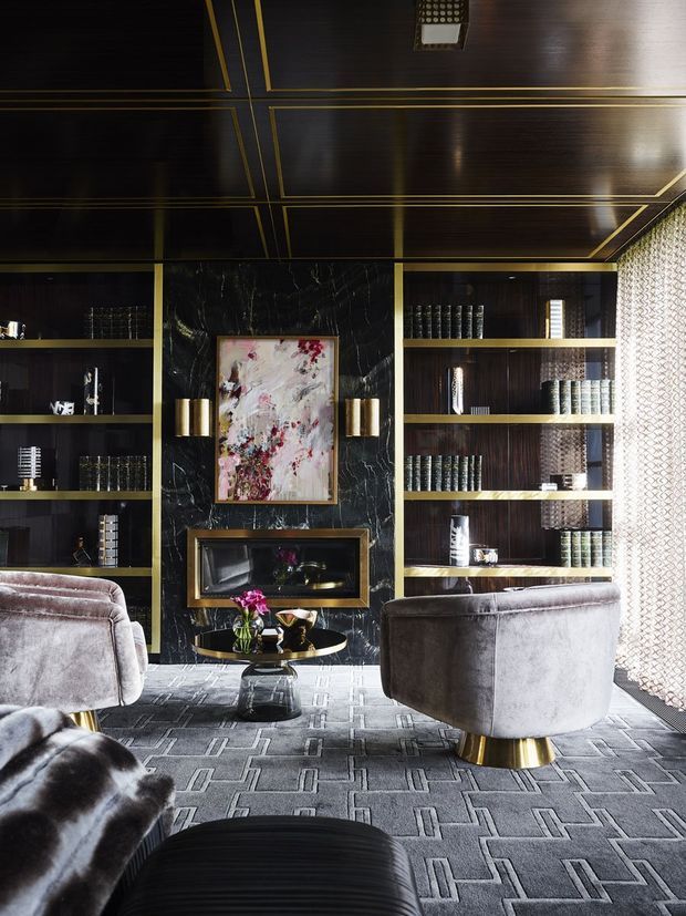 A sophisticated, dark-toned living room or lounge with floor-to-ceiling built-in shelving, a black marble fireplace surround, brass detailing, and a geometric patterned ceiling. Plush grey velvet lounge chairs face the fireplace, with a round black coffee table and decorative artwork. A patterned sheer curtain filters daylight at the right, while the room is grounded by a grey geometric carpet.