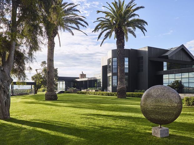 A contemporary dark-clad building set within a manicured lawn, framed by mature palm trees and landscaped hedges. The complex features large glazed openings, strong geometric volumes, and a prominent spherical metal sculpture on a plinth in the foreground.