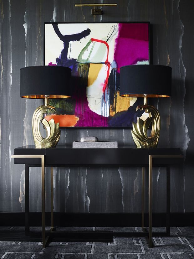 A dramatic, symmetrical interior vignette featuring a slim black console table with brass-toned legs, two sculptural metallic table lamps with black drum shades, and a colourful abstract artwork. The backdrop is a dark vertically textured wall, with a dark patterned carpet underfoot. The styling is bold, luxurious, and highly contrasted.