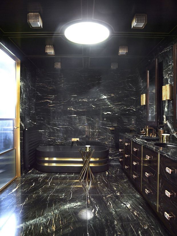 Luxurious, dramatically lit black marble bathroom with matching marble floor and walls, a freestanding oval bathtub with metallic trim, dark custom vanity cabinetry, large mirror, brass or gold-toned fixtures, and geometric pendant-style ceiling lights. The space has a high-contrast, glamorous art deco character.