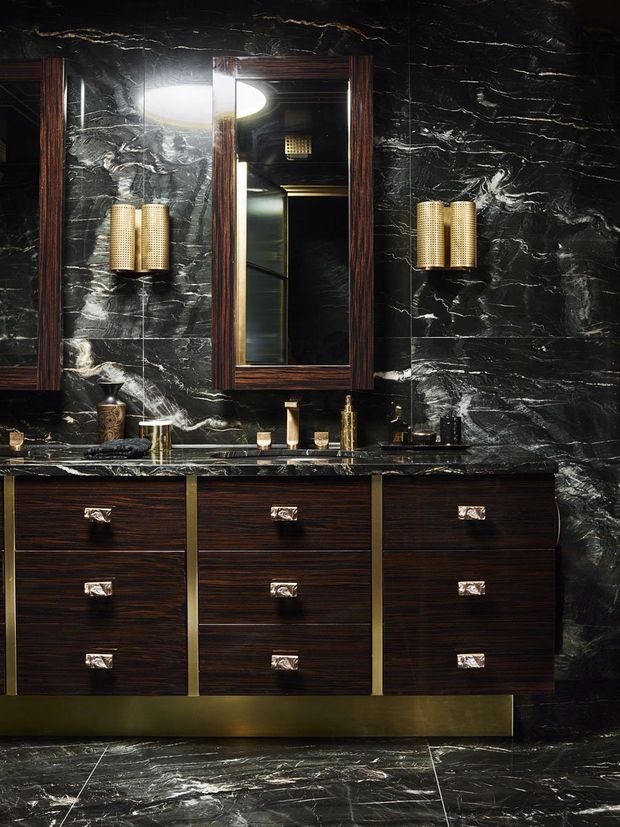 A dramatic, luxurious bathroom vanity with dark timber cabinetry, a black-and-white veined stone wall and matching stone benchtop, brass-toned wall sconces and hardware, a central framed mirror, and dark veined stone flooring. The space has a moody Art Deco-inspired aesthetic with rich materials and high contrast.