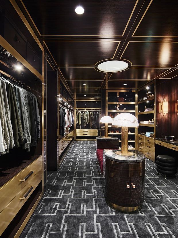 Luxurious walk-in wardrobe and dressing room with dark timber wall and ceiling panelling outlined in brass-toned trim. The room features extensive open clothing rails, mirrored cabinetry, brass and dark wood drawers, patterned dark carpet, a central cylindrical island with a marble top and sculptural lighting, plus a red upholstered bench and black ottoman. Dramatic layered lighting creates a refined, high-contrast atmosphere.