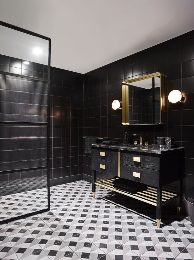 A dramatic contemporary bathroom with black tiled walls, a geometric black-and-white tiled floor, a glass shower enclosure, and a freestanding dark vanity with brass detailing. A rounded-rectangle brass-framed mirror and globe wall lights provide warm metallic accents.