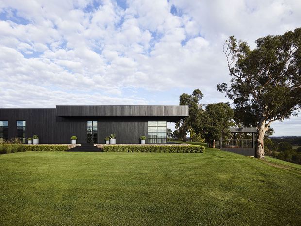 Exterior view of a contemporary single-storey home set on a broad landscaped lawn. The building has a dark vertically textured facade, a strong horizontal flat roofline, extensive rectangular glazing, and a covered open-sided area at the right. Mature trees and a distant elevated landscape surround the property.