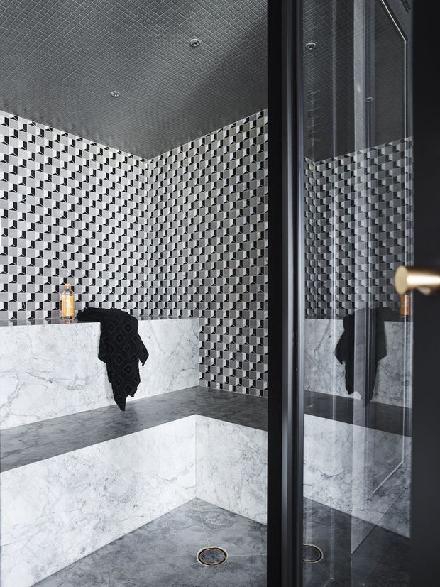 A dramatic monochrome bathroom or shower room clad in geometric black, white and grey mosaic wall tiles. Large white veined marble slabs form built-in bench seating and lower wall surfaces, complemented by grey stone-look flooring and a dark textured ceiling. A frameless or dark-framed glass shower enclosure and reflections are visible on the right, with a black towel draped over the bench.
