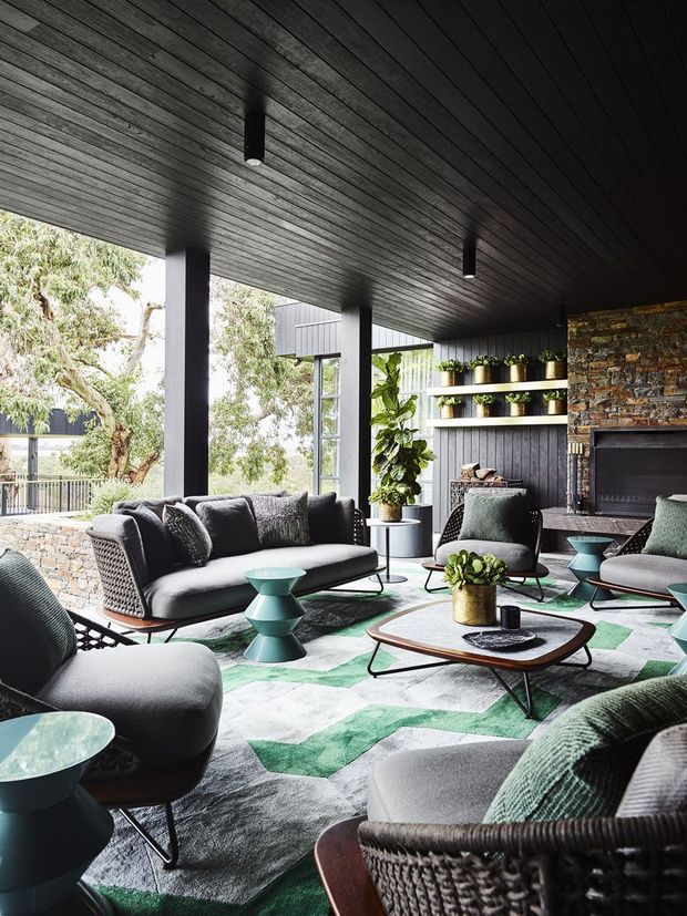 A sophisticated indoor-outdoor living room with extensive glazing opening to a leafy setting. The room features a dark timber-lined ceiling, charcoal vertical wall panelling, black structural columns, a stacked natural-stone fireplace wall, open shelving with plants, and a coordinated arrangement of grey upholstered sofas and woven lounge chairs around low mid-century-style coffee tables. A large geometric grey, white, and green rug and teal accent tables add colour and pattern.