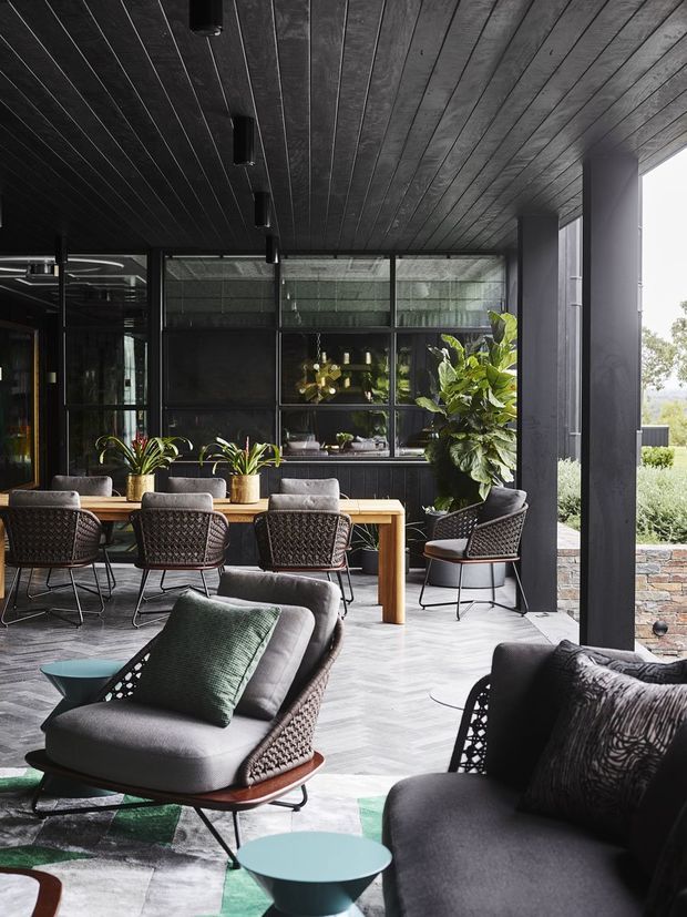 A sophisticated covered outdoor entertaining area with a dining table, woven lounge chairs, and low sofas arranged on a pale stone or concrete floor. Dark timber-lined ceilings, black-framed glazing, structural columns, and extensive indoor plants create a dramatic indoor-outdoor setting. A stone-clad low wall and landscaped surroundings are visible beyond the open edge.