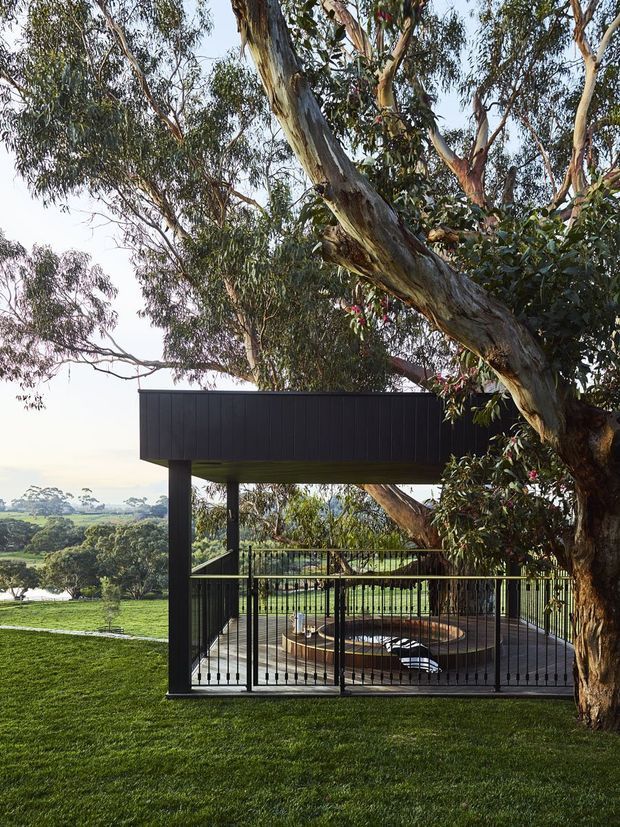 A contemporary open-air pavilion or outdoor lounge set within a landscaped garden and mature eucalyptus trees. The structure has dark vertical cladding, slender black structural posts and railings, and a timber deck surrounding a circular recessed fire pit or seating feature. The elevated site overlooks a broad green landscape and distant trees.