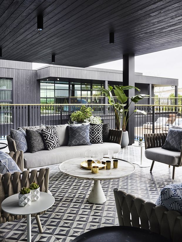 A sophisticated covered outdoor lounge or terrace with a contemporary black-and-white scheme. The space features a dark timber-lined ceiling with recessed pendant lights, black structural posts and railings, patterned geometric floor tiles, a white sofa and armchairs with bold monochrome cushions, and a round marble-look coffee table. Large glazed openings connect to the modern building beyond, while potted tropical plants add greenery.