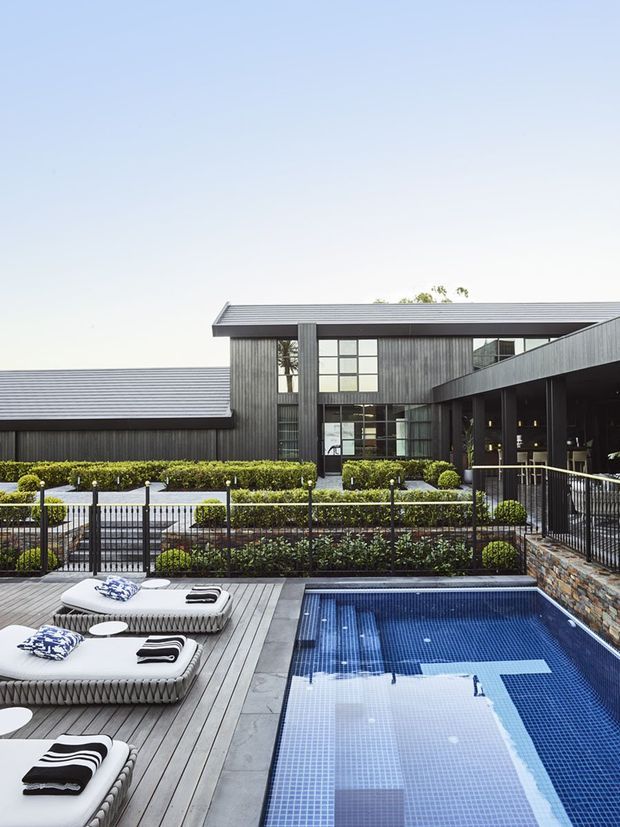 A modern dark-clad building viewed from an outdoor pool terrace. The rectangular swimming pool is bordered by grey timber decking, sculptural white loungers, manicured hedges, black metal fencing, and a stone-clad retaining wall. The building has charcoal vertical cladding, low-pitched metal roofs, large grid-patterned windows, and a covered outdoor walkway or terrace.