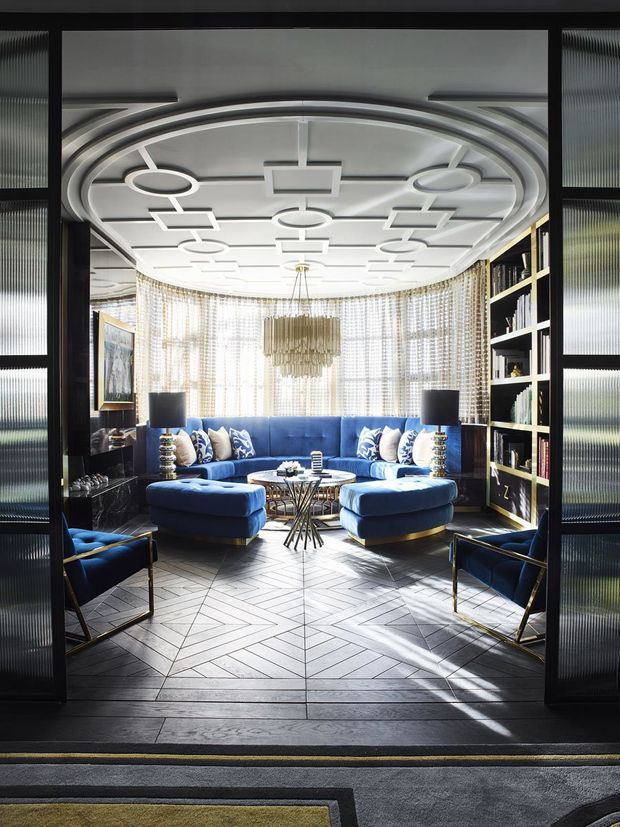 A highly styled, symmetrical living room viewed through dark-framed glass doors. The room features a vivid blue curved sectional sofa, matching blue lounge chairs, patterned cushions, black built-in shelving, metallic side lamps, a sculptural chandelier, and an elaborate white coffered ceiling with concentric curved detailing. Floor-to-ceiling sheer patterned curtains diffuse abundant daylight, while geometric timber flooring and dark thresholds frame the space.