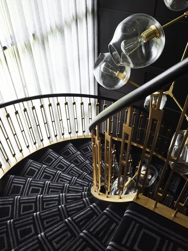 A dramatic curved stairway with black patterned carpet, dark wall panelling, and ornate gold geometric balustrades. Sculptural glass globe pendant lights with brass fittings hang alongside the stair, creating a luxurious Art Deco-inspired interior.