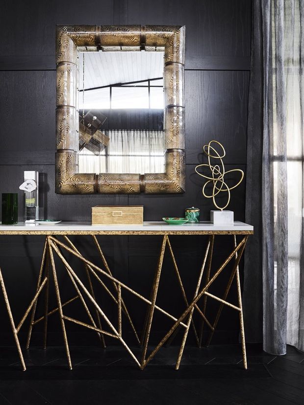 A dramatic, highly styled interior vignette featuring a narrow console table with an angular gold metal frame and pale stone or marble top, set against a black vertically panelled wall. Above it hangs a large ornate metallic gold-framed mirror. Small decorative objects sit on the console, while sheer floor-length curtains frame the right side and reflect the bright window beyond. The composition has a luxurious, sculptural, high-contrast character.