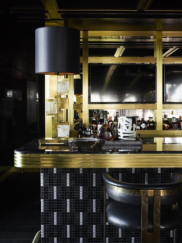 A dramatic, highly styled home bar with a black tiled counter front, glossy dark surfaces, brass-gold trim and framing, illuminated mirrored shelving, bottles, glassware, and a black leather bar stool. The space has strong Art Deco-inspired detailing, including rounded counter edges, metallic ribbed accents, geometric tile patterning, and a large cylindrical pendant light.