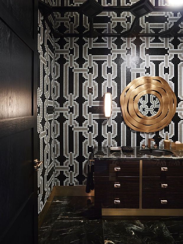 A dramatic, compact bathroom or powder room featuring a bold black-and-white geometric mosaic tile pattern covering the walls and ceiling. A dark timber vanity with multiple drawers supports a dark stone benchtop and integrated basin, while a large concentric brass-gold circular mirror and warm metallic details provide a luxurious focal point. The room has glossy dark marble or stone flooring, black timber doors and joinery, and moody directional lighting.