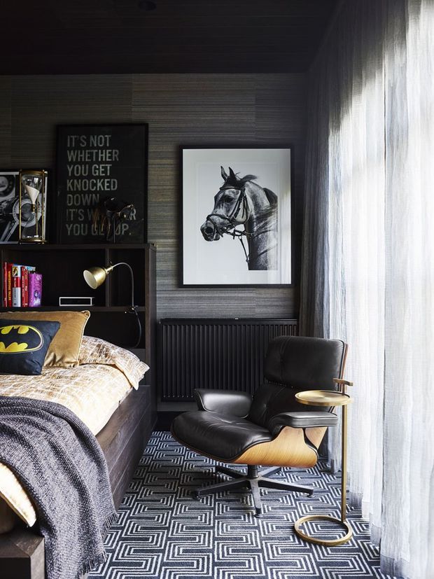 A moody, compact bedroom with dark timber ceiling and built-in joinery, textured grey wall panelling, a black radiator, and a large sheer-curtained window. The room features a timber bed with patterned bedding, a black leather lounge chair with an integrated side table, geometric black-and-white carpet, books, framed horse artwork, and a motivational wall print.