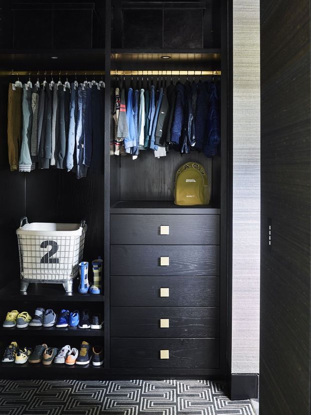 A dark, highly organised walk-in wardrobe with black wood-grain built-in cabinetry, open hanging rails, multiple drawers, shoe shelving, and integrated lighting. Clothes, shoes, and a laundry basket are stored within the joinery, with a bold geometric carpet covering the floor.