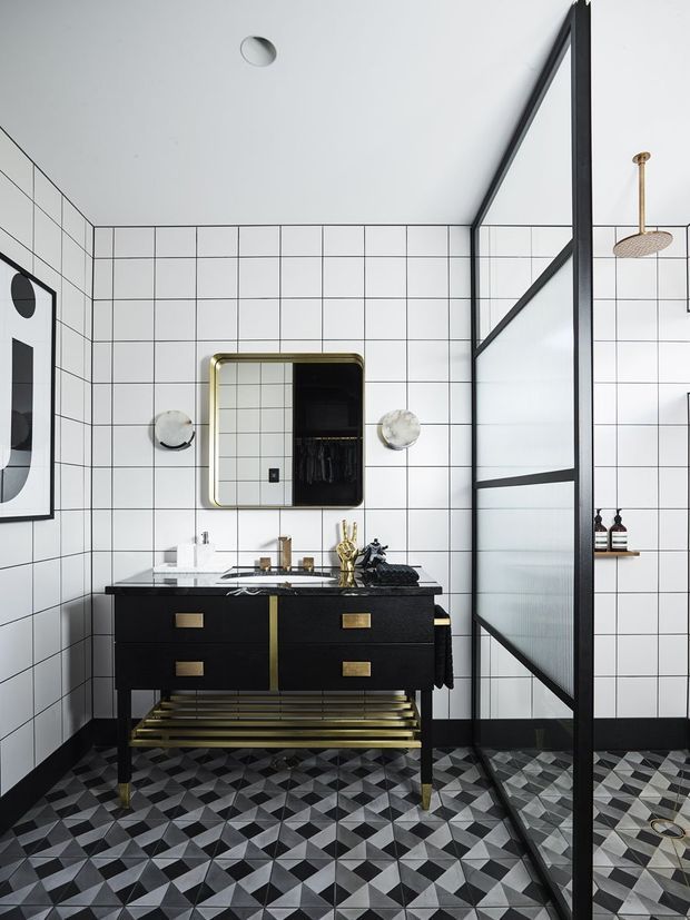 A striking contemporary bathroom featuring white square grid wall tiles, a geometric black, white and grey patterned floor, and a black double vanity with brass hardware and legs. A rounded rectangular brass-framed mirror is mounted above the vanity, with brass wall lights and fixtures. A large black-framed frosted-glass shower screen occupies the right side, with an exposed brass showerhead and wall shelf holding toiletries. The room has a bold monochrome palette with warm metallic accents and strong graphic detailing.