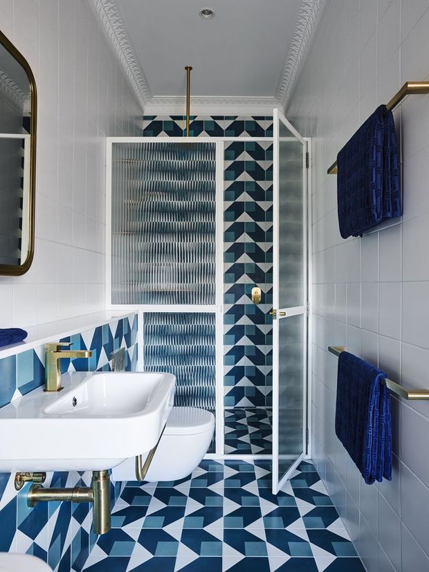 Compact, highly styled bathroom with a white wall-tiled envelope, ornate decorative ceiling cornices, and bold geometric blue, teal, black, and white patterned tiles running across the shower wall, floor, and lower vanity wall. A white wall-mounted basin with exposed brass-toned plumbing sits in the foreground, with a concealed toilet behind it. The walk-in shower has a clear framed glass door and patterned tile interior. Brass-toned fixtures, towel rails, and a wall-mounted navy towel provide refined contrast. A rounded rectangular mirror is partially visible on the left.