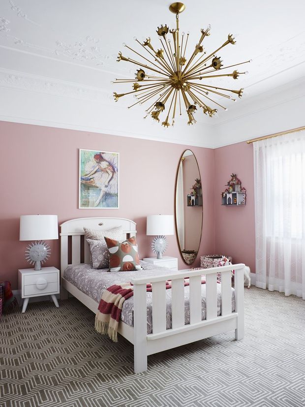 A stylish bedroom with blush pink walls, ornate white ceiling cornices, and a dramatic brass starburst chandelier. The room features a white spindle-style bed with patterned bedding, matching white bedside tables and lamps, a tall oval mirror, small wall shelves, sheer curtains, and a bold grey-and-white geometric carpet. The look is playful, eclectic, and refined with a strong mid-century-inspired lighting feature.