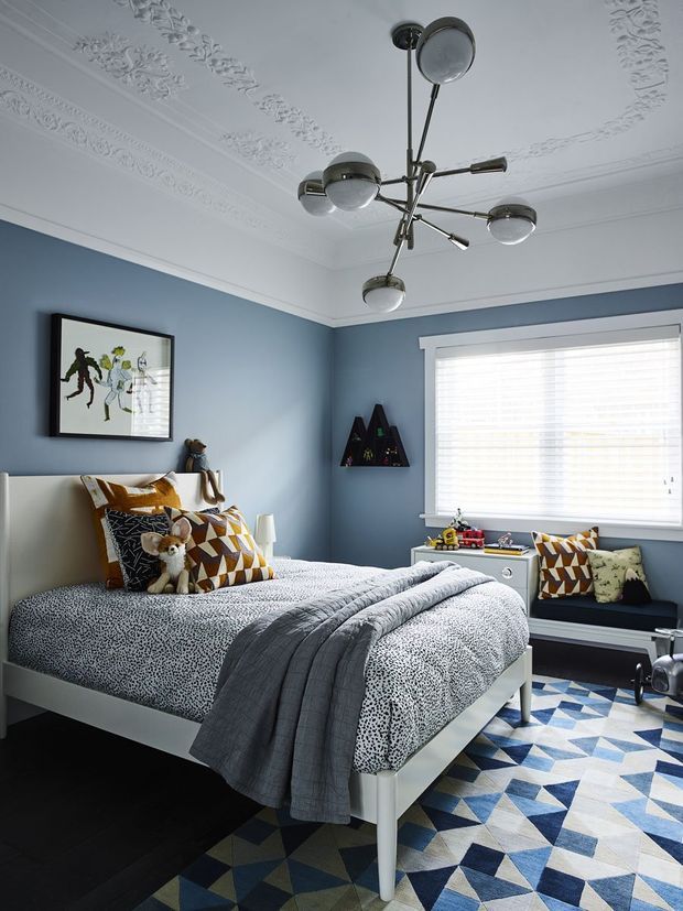 A stylish children's bedroom with blue walls, an ornate white decorative ceiling, and a modern multi-arm spherical pendant light. The room features a white bed with patterned bedding and geometric accent pillows, a dark upholstered window seat, framed artwork, small wall shelving, and a blue, white, and grey geometric area rug. Natural light enters through a large window with white blinds.