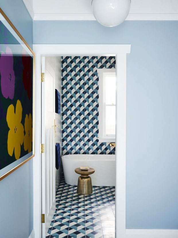 View from a light blue hallway into a boldly patterned bathroom. The bathroom features a freestanding white bathtub, geometric blue, white and dark patterned tile covering the floor and rear wall, a white-framed window, and a small metallic gold stool or side table. The hallway has white trim, a spherical pendant light, and a large colourful framed artwork.