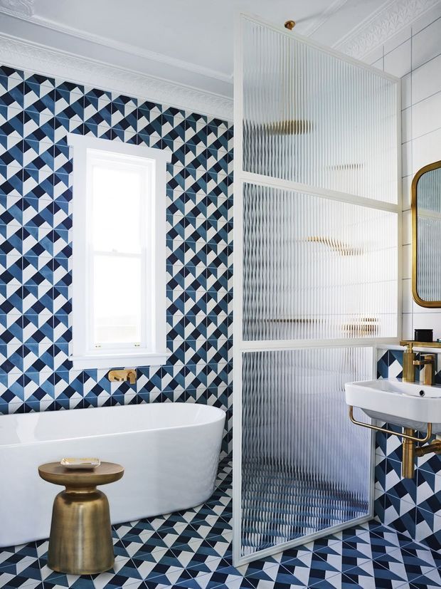 A highly patterned bathroom featuring a freestanding white bathtub, wall-mounted white basin with brass plumbing, brass-framed mirror, and a ribbed translucent glass partition. Blue, navy, white, and pale pink geometric tiles cover the walls and floor, complemented by ornate white ceiling mouldings and brass fixtures.