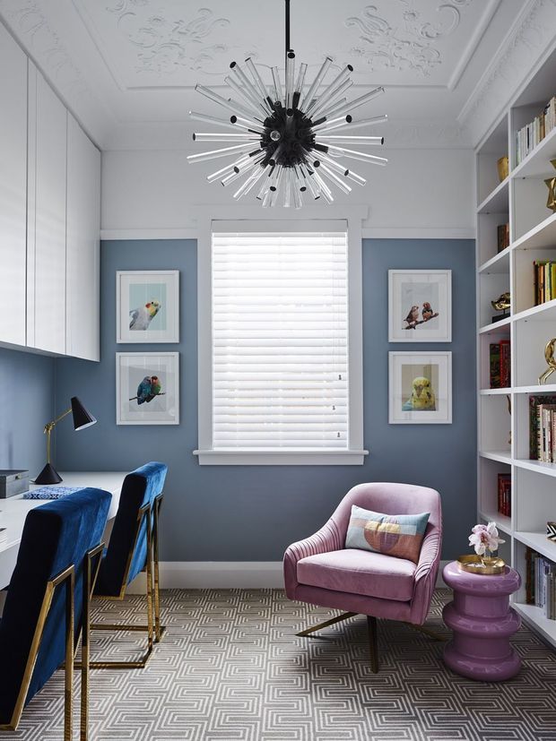 A refined, compact home office with dusty blue walls, white built-in cabinetry and open shelving, a large window with white plantation shutters, and ornate white decorative ceiling panels. The room features a striking mid-century-inspired starburst pendant, two blue upholstered desk chairs at a white work surface, a mauve-pink lounge chair, a glossy purple side table, framed bird artwork, books, and decorative objects. A patterned geometric carpet adds visual interest, creating a colourful, elegant and eclectic workspace.