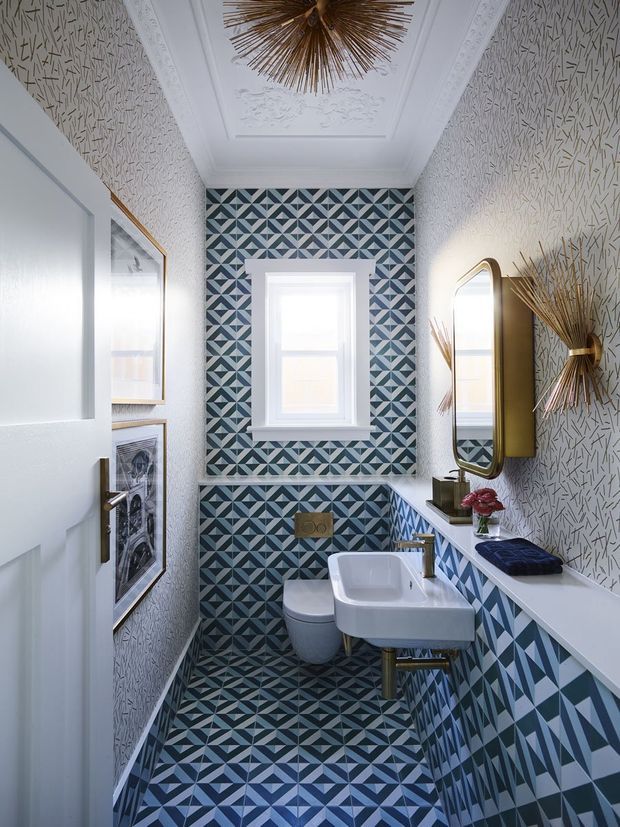 A narrow, highly decorative bathroom with bold geometric blue, grey and white patterned tiles wrapping the floor, lower walls and window wall. White textured wallpaper covers the upper walls, while the ceiling features ornate plaster detailing and a sculptural brass starburst light. The room includes a wall-mounted white basin with brass tapware, a compact toilet, a framed mirror, brass wall lighting and a small windowsill with floral decor. The overall effect is richly patterned, refined and eclectic with strong Art Deco influences.