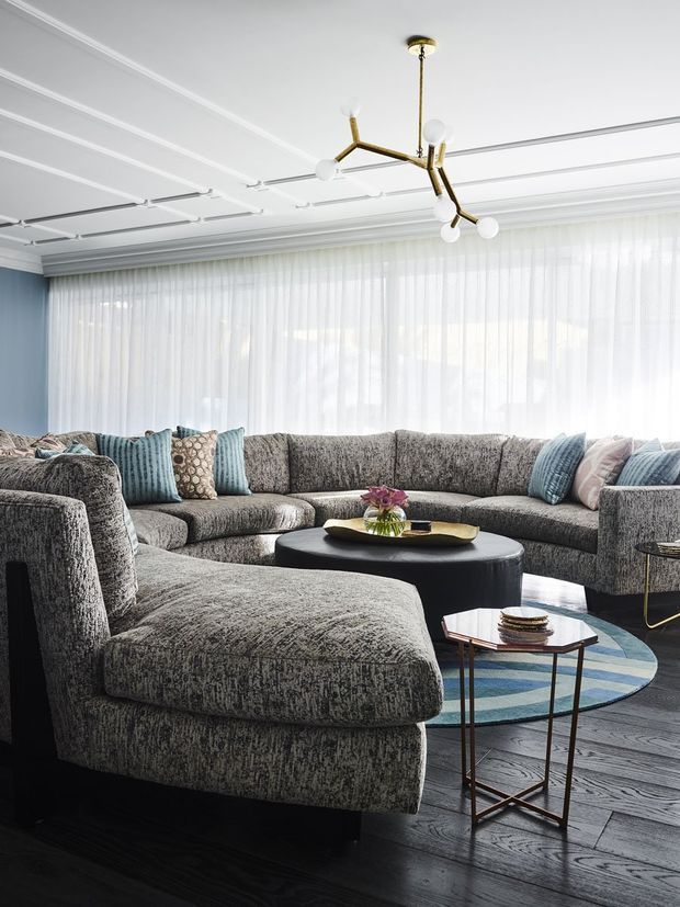 Bright, refined living room with a large curved grey sectional sofa arranged around a low black oval coffee table. A patterned blue and grey rug, assorted cushions, side tables, and floral centrepiece add layered furnishings. Floor-to-ceiling sheer curtains span the rear wall, while the white coffered and panelled ceiling features a sculptural brass multi-arm pendant light. Dark timber flooring and a pale blue wall create a sophisticated, contemporary interior.