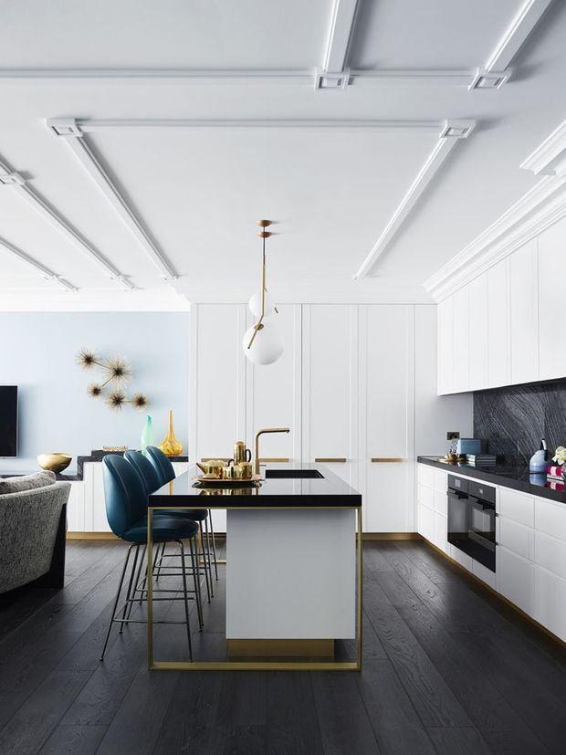 A bright, refined contemporary kitchen with white full-height cabinetry, a large island with a white waterfall-style front and black benchtop, and integrated black appliances. Deep charcoal timber flooring contrasts with the white joinery, while brass hardware, tapware and island detailing add warmth. Blue upholstered bar stools sit at the island, and a sculptural pendant hangs beneath a decorative white ceiling with linear mouldings. The adjoining living area is partially visible at left.