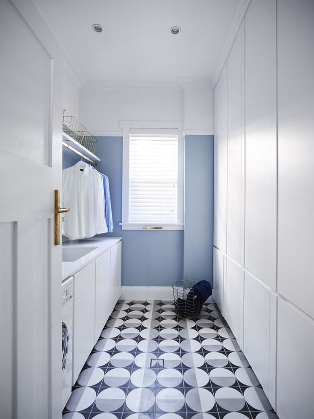 A narrow, bright laundry room with white built-in cabinetry and a white benchtop along the left side, integrated washing appliances, open hanging rails with white and pale blue clothing, and full-height storage cupboards on the right. The room has pale blue walls, white trim and ceiling cornices, recessed downlights, a central shuttered window, and a bold geometric black, white and grey patterned tile floor. A basket and folded textiles sit near the far end.