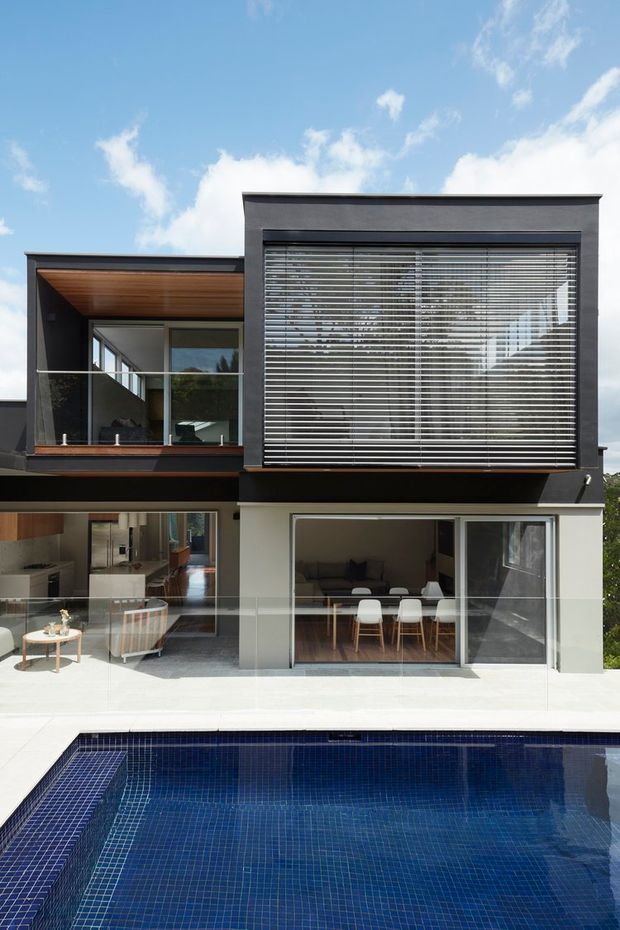Exterior view of a contemporary multi-level residence with dark charcoal cladding, extensive glazing, timber soffits, and horizontal external blinds. A glass-balustraded terrace overlooks a deep blue tiled swimming pool, with living and dining areas visible through the ground-floor openings.
