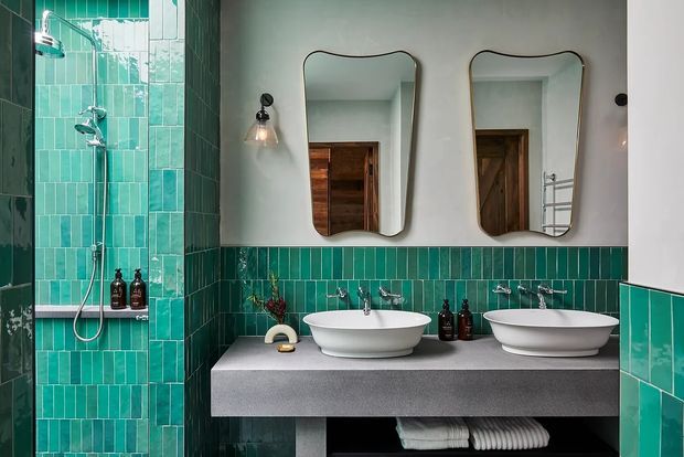 Contemporary double-vanity bathroom with a walk-in shower, vivid turquoise-green vertical wall tiles, a grey concrete-look vanity top, two white vessel basins, organic-shaped mirrors, black wall sconces and dark timber reflected in the mirrors.
