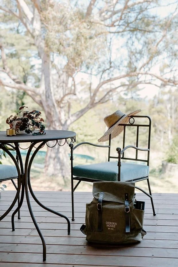 A tranquil covered outdoor deck or veranda furnished with a dark metal café-style table and matching metal-framed chairs with blue-green cushions. A potted plant sits on the table, while a canvas backpack rests on the timber decking. Large glazing or an open edge looks out onto leafy trees and a softly visible blue-green pool or landscape beyond. The scene has a relaxed, vintage outdoor-living character.