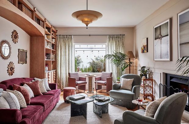 A refined traditional living room with a burgundy sofa, upholstered armchairs, marble coffee tables, built-in timber shelving, decorative wall mouldings, a fireplace, patterned curtains, and indoor palms. A bright window seating area sits at the rear, while warm layered lighting and rich textiles create an inviting, sophisticated atmosphere.